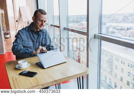 Successful Happy Business Man Is Sitting At A Table In A Cafe, Holding A Cup Of Coffee And Using A L