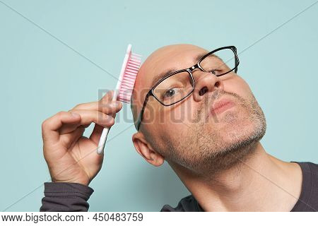 Bald, Bearded Man With Glasses And A Funny Face Looks Into The Camera And Combs With A Baby Comb, On