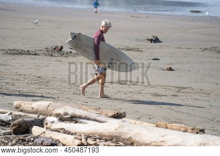 Tolaga Bay - New Zealand - February 5 2022; Lone Surfer Walks Along Beach Carrying Surfboard To Wate