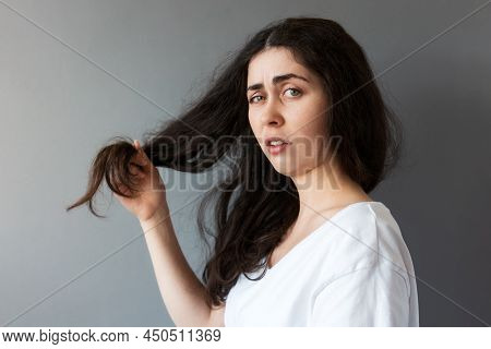 Portrait Of Young Upset Woman Show Dry Tips Of Her Long Dark Hair. Gray Background. The Concept Of T