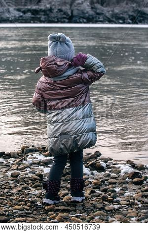 A Little Girl On A Winter Walk Stands With Her Back To Us And Throws Stones Into A Cold River