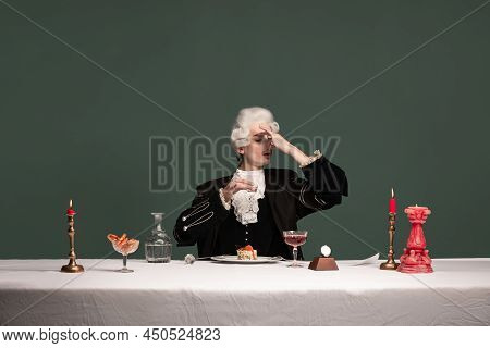 Portrait Of Young Elegant Man In Peruke And Vintage Jacket Sitting At Table Isolated On Dark Green B