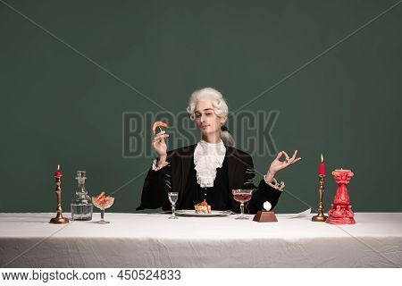 Portrait Of Young Elegant Man In Peruke And Vintage Jacket Sitting At Table Isolated On Dark Green B