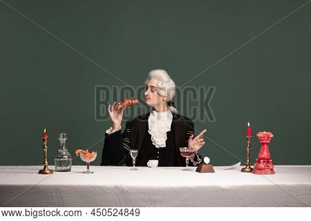 Portrait Of Young Elegant Man In Peruke And Vintage Jacket Sitting At Table Isolated On Dark Green B
