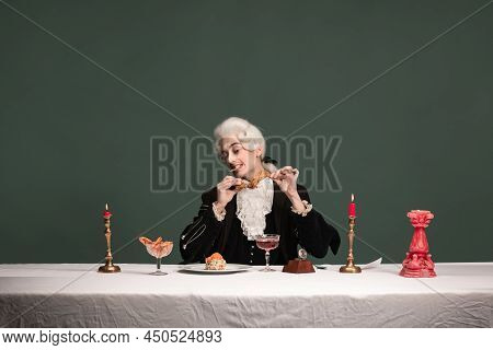 Portrait Of Young Elegant Man In Peruke And Vintage Jacket Sitting At Table Isolated On Dark Green B