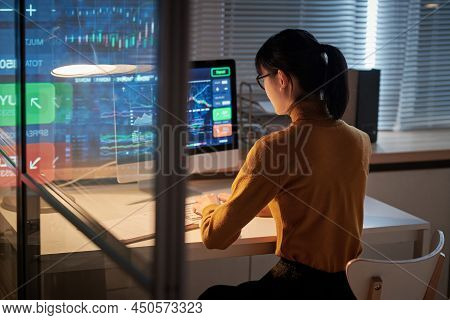 Rear View Of Young Woman Sitting At Table In Front Of Computer Monitor And Working With Software At 