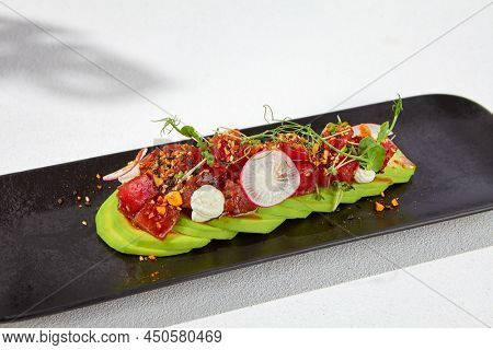 Modern food - tuna tartare on sliced avocado in black plate. Avocado starter with tuna and radish in asian style. Contemporary asian dish. Healthy food with avo and marinated fish on white background