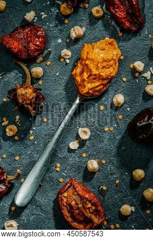 high angle view of a spoon with some romesco sauce, typical of Catalonia, Spain, on a dark surface with some of the ingredients to prepare it, such as tomatoes, garlic, nyora pepper or hazelnuts