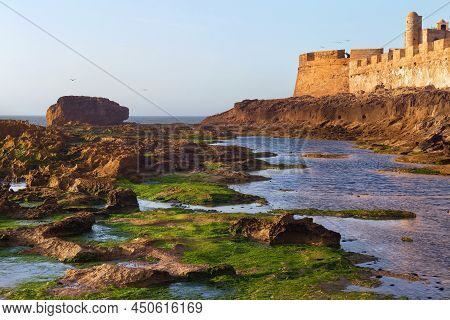 View Of The Historical Walls Of The Essaouira Fortress And Volcanic Shore Of The Atlantic Ocean In M