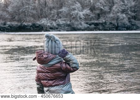 A Little Girl On A Winter Walk Stands With Her Back To Us And Throws Stones Into A Cold River