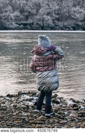 A Little Girl On A Winter Walk Stands With Her Back To Us And Throws Stones Into A Cold River