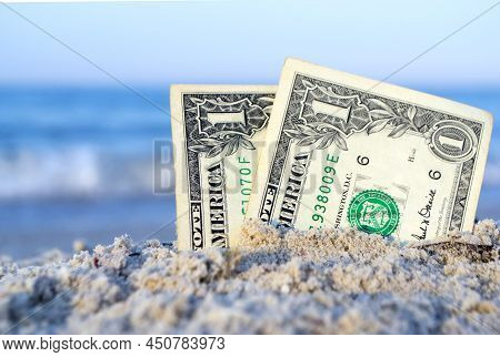 Two One Dollar Bills Half Buried In Sand On Sandy Seashore Close-up.