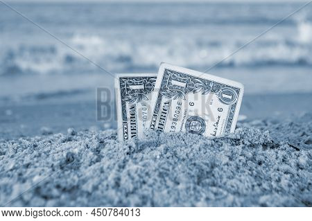 Two One Dollar Bills Half Buried In Sand On Sandy Seashore Close-up.
