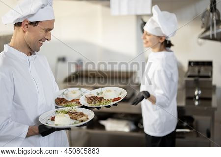 Chef Standing With Ready Meals, Talking With Female Cook In The Kitchen. Happy Caucasian Cook Workin