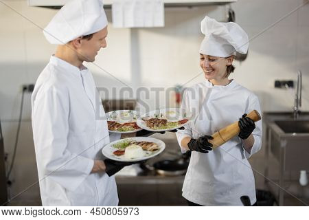 Chef Standing With Ready Meals, Talking With Female Cook In The Kitchen. Happy Caucasian Cook Workin