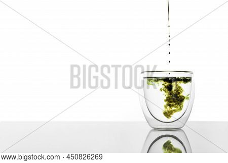Glass Of Water With Chlorophyll Extract Against White Grey Background. Pouring Liquid Chlorophyll In