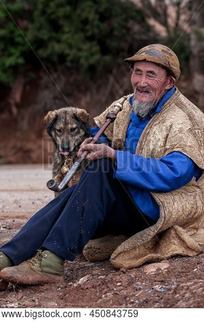 Yunnan, China - March 5, 2012: Portrait Of An Elderly Chinese Bearded Man With Dog Smiling While Smo