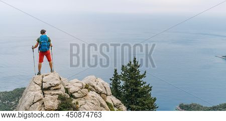 Young Hiker Backpacker Man Using Trekking Poles Enjoying The Mediterranean Sea On Clifftop During Ly