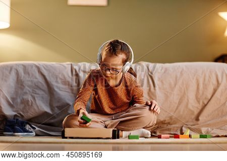 Portrait Of Young Girl With Down Syndrome Playing With Toy Blocks Alone And Wearing Noise Cancelling