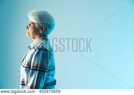 Sad And Pensive Older Gray-hared Lady Standing Profile. Medium Studio Shot Over Blue Background. Hig