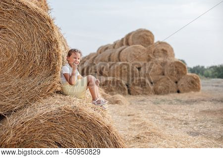 Excited Little Girl Sitting On Rolled Haystack In Sundress Resting Back On Another Hayrick Picking T