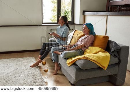 Side View Two Joyful Barefoot Lesbian Women Sitting On Sofa, Playing Video Games. Two Glasses Of Win
