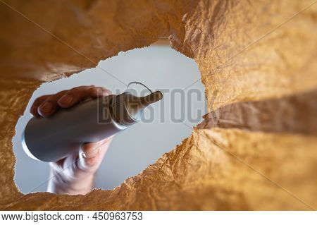 A Hand Places A Gray Plastic Bottle In A Paper Bag. A Mature Man's Hand Holds A Sweet Ice Cream Topp