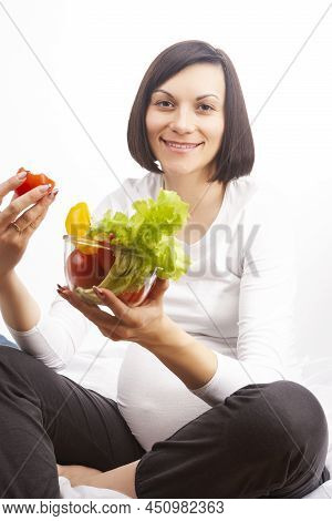 Young Pregnant Brunette Woman Sitting On Fitball And Eating Fresh Vegetables In Glass Bowl Over Whit