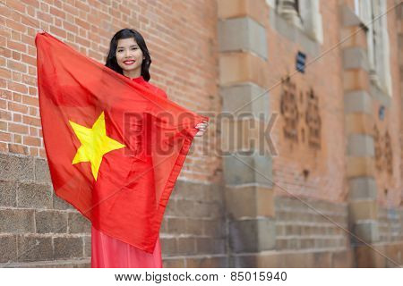 Happy patriotic young Vietnamese woman with a lovely warm smile holding up the national flag in her arms as she stands against brick buildings in an urban street, with copyspace