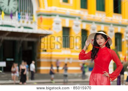 Stylish young Vietnamese woman in red standing raising her hand elegantly to her traditional hat as she stands in an urban street with colorful buildings