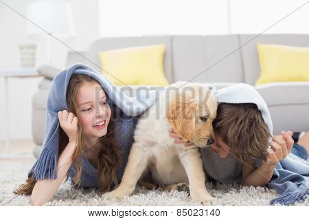 Brother and sister with puppy under blanket lying on rug at home