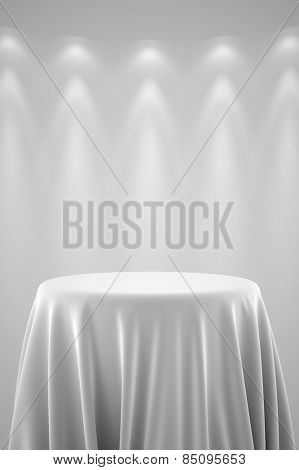 Round presentation pedestal covered with a white silk cloth in front of a wall illuminated by spot lights