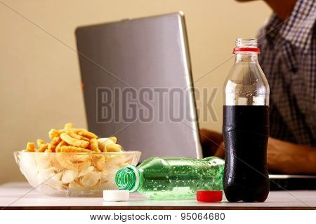 Bottles of softdrinks or soda, chips and man working on a laptop computer in the background