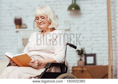 Time for hobby. Pleasant delighted elderly woman in wheelchair reading book while resting at home
