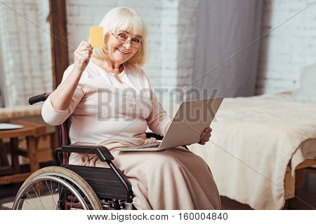 Online service. Cheerful elderly disabled woman holding credit card and using laptop while sitting in the wheelchair