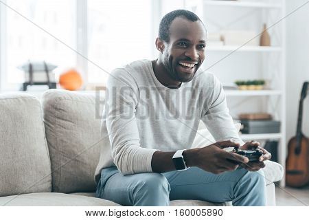 Resting at home. Handsome young African man playing video games and laughing while sitting on the sofa at home