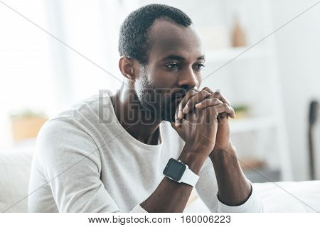 Something on his mind. Handsome young African man holding hands together and thinking while sitting on the sofa at home