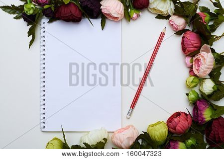 A mid shot of white jotter and red pencil circled by beautiful peonies lying on the light field. Excellent image of stylish objects