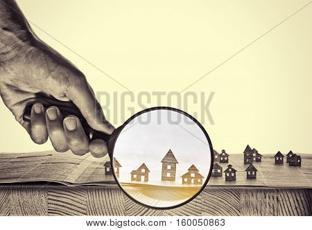 Hand holds the magnifying glass in front of an open newspaper with paper houses. That could mean rent search purchase real estate.