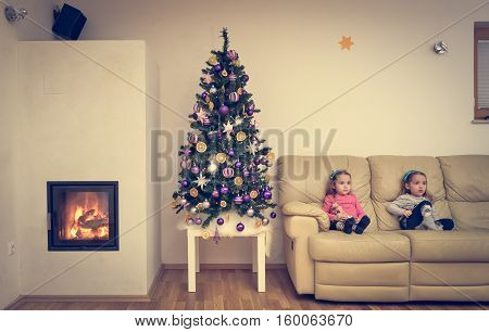 Children and Christmas tree in modern luxury apartment with fireplace. Twin girls are sitting on a sofa next to home decorated Christmas tree with hot fire and flames burning in the fireplace.