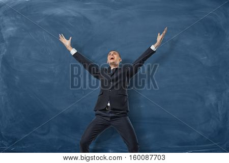 A businessman standing with splayed arms and shouting on the blue blackboard background. Bright and vivid emotions. The best in his field. Business development and the way to success. With great enthusiasm