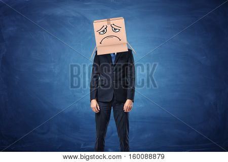 Businessman wearing cardboard box with drawn sad face on his head near the blue blackboard. Anonymous person. Packing and crating. Issues of small business.