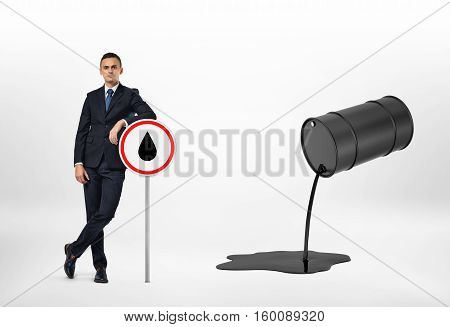 A businessman leaning on the road sign with the oil drop depicted on it, and oil pouring out of the black barrel, isolated on the white background. Oil and petroleum products. Black gold. Resource extraction.