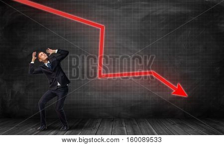 A small frightened businessman under a huge red kinked arrow pointing down on a dark plaid background. Business falling down. Negative trend. Crisis impact.