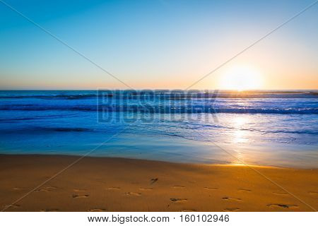 Sand beach with traces of footprints and ocean surf is painted in the colors of the setting sun. Coast of California.