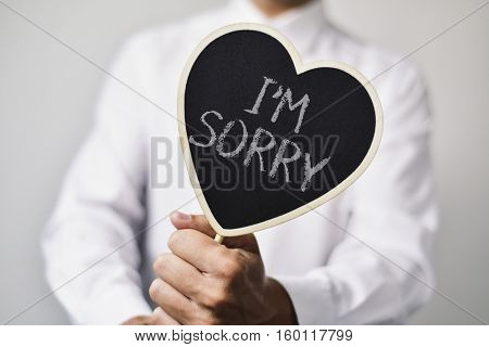 a young caucasian man wearing a white shirt shows a heart-shaped signboard with the text I am sorry written in it
