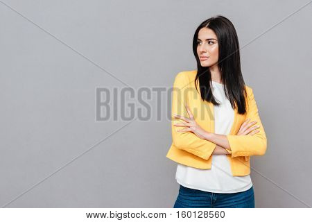 Picture of attractive young woman dressed in yellow jacket posing over grey background with arms crossed while looking at copyspace.