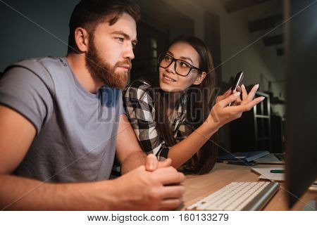 Two business colleagues working late at night in their office with computer. Woman talking to man. Man looking at computer.