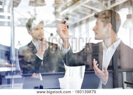 Two concetrated young businessmen talking and writing on glass board in office