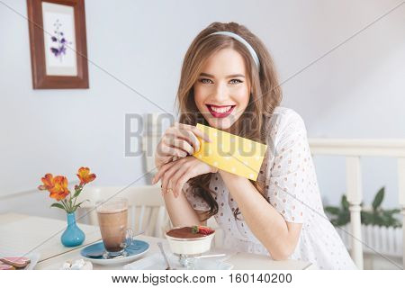Cheeerful attractive young woman in white dress drinking latte and eating dessert in cafe